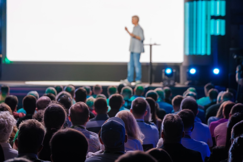 A speaker presents on stage to a large audience, illuminated by colorful lights, with a blank screen behind them.