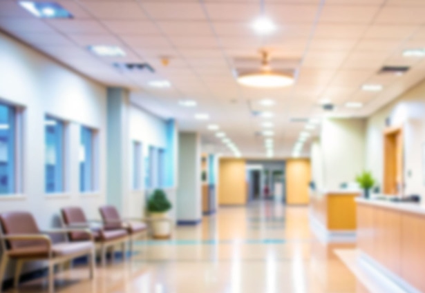Blurred view of a bright hospital hallway with waiting chairs, plants, and a reception desk in a modern, clean environment.